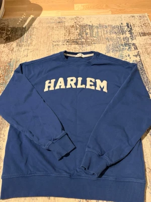 Blå sweatshirt HARLEM från MNG - Blå sweatshirt från MNG med vit HARLEM-tryck på bröstet och text på ryggen. Tröjan har rund hals, ribbade muddar vid ärmslut och nederkant samt långärmad passform. Perfekt för chill dagar och streetstyle.