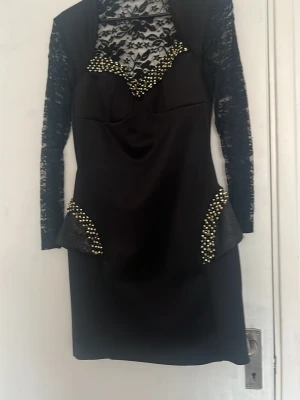 Svart kort klänning med spets och pärlor - Snygg svart kort klänning med långa spetsärmar och spetsdetaljer vid halsen. Dekorerad med pärlor vid halsringningen och på sidorna. Klänningen har en figurnära passform och coola detaljer som ger en unik look.