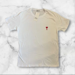 Vit t-shirt från Ami med hjärtdetalj - Säljer en fräsch vit t-shirt från Ami med en liten röd hjärtdetalj och bokstaven A broderad på bröstet. T-shirten har rund hals och korta ärmar, perfekt för dig som gillar stilrena plagg med en twist.