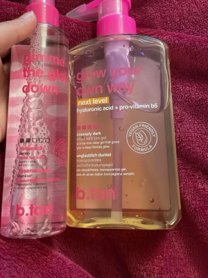 b.tan glow self tan gel & mist - Säljer två produkter från b.tan: 'gimme the glow down' facial tan mist med rosenvatten i en genomskinlig plastflaska med rosa text och pump, samt 'glow your own way' clear self tan gel med hyaluronsyra och pro-vitamin B5 i en större pumpflaska. Båda är veganska och ger en bronzig glow.