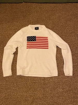 Vit stickad tröja med USA-flagga - Vit stickad tröja med en stor amerikansk flagga på bröstet. Tröjan har rund halsringning och långa ärmar. Perfekt för dig som vill ha en klassisk och stilren look med en twist.