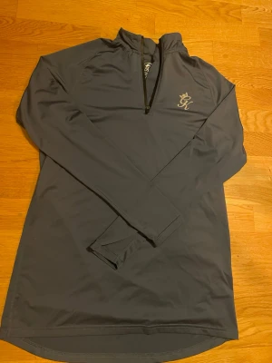 Grå långärmad träningströja Gymking - Snygg grå långärmad träningströja från Gymking med halv dragkedja och diskret logga på bröstet. Tillverkad i mjukt och stretchigt syntetmaterial som andas, perfekt för gymmet eller löprundan. Tröjan har en stilren design och bekväm passform.