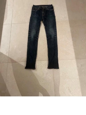Mörkblå Nudie Jeans Skinny - Säljer ett par mörkblå skinny jeans från Nudie Jeans med klassiska fem fickor och snygga slitningar vid knäna. Jeansen har orangea sömmar och Nudie-logga på bakfickan. Perfekt passform med låg midja och stretchigt denim-material.