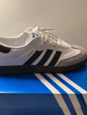 Adidas Samba sneakers vit/svart - Adidas Samba sneakers med klassisk design i vitt och svart, med grå mockadetaljer på tån. Skorna har snörning, platt sula och de ikoniska tre ränderna på sidan. Märket syns på tungan och sidan. Perfekt för dig som gillar retrostil och streetwear.