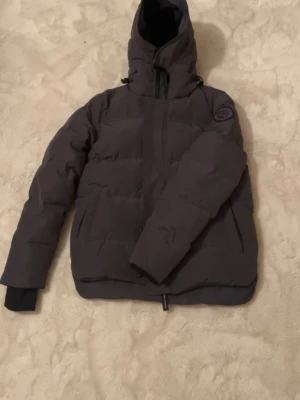 Canada Goose jacka - Svart dunjacka från Canada Goose med huva och diskret logga på ärmen. Jackan har quiltad design, dragkedja framtill och ribbade muddar vid ärmslut. Perfekt för kalla dagar och har en clean, modern look som funkar till streetwear.