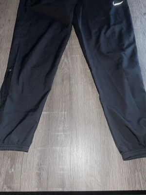 Svarta träningsbyxor från Nike - Svarta träningsbyxor från Nike med elastiska benslut och dragkedja vid anklarna. Byxorna har en stilren design med Nike-logga på höften och är gjorda i lätt polyester som passar perfekt för sport och aktivitet.