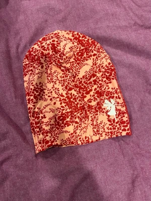 Röd blommig beanie House of Lola - Snygg bomullsbeanie med rött blommigt mönster på ljus bakgrund och en liten vit broderad figur på sidan (House of Lola). Perfekt för dig som vill sticka ut med en färgglad accessoar. Mjuk och skön passform.