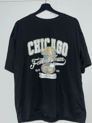 Svart t-shirt - Svart t-shirt med stort tryck 'CHICAGO Football Team' i vitt på framsidan. T-shirten har korta ärmar och en klassisk rund hals. Använd flertal gång och trycket har börjat flagna. Från SHEIN 