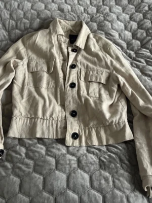 Beige linnejacka från New Look - Snygg beige linnejacka från New Look med stora svarta knappar och två bröstfickor. Jackan har klassisk krage och är tillverkad i ett linneliknande material som ger en chill vibe. Perfekt att slänga på över en t-shirt för en laidback look.