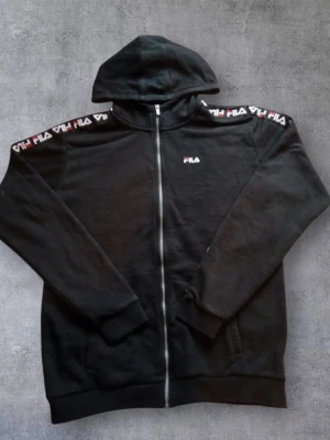 FILA hoodie / huvtröja svart – stl 170–176 - Svart FILA hoodie med dragkedja och huva. Logga på bröstet och längs ärmarna. Storlek: 170–176 cm (ca 15–16 år). I fint begagnat skick, inga hål eller fläckar. Perfekt till vardag.