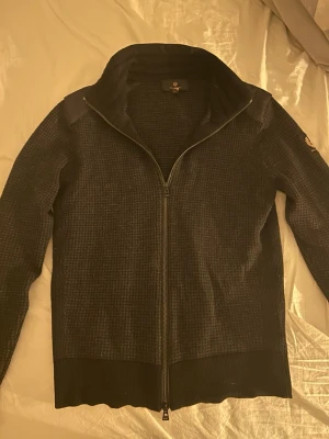 Svart kofta från Belstaff med dragkedja - Säljer en svart kofta från Belstaff i storlek XS. Koftan har ett diskret rutigt mönster, hel dragkedja framtill och ribbade muddar. Materialet är stickat och känns mjukt och bekvämt. Snygg logga på ärmen och förstärkta axelpartier.  Nypris: 3500 köpt på NK