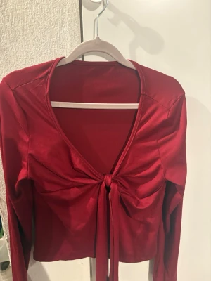 Röd omlottopp med knyt framtill - Säljer en snygg röd omlottopp med lång ärm och v-ringning. Toppen har ett knyt framtill som ger en cool och trendig look. Perfekt att styla med jeans eller kjol för en chic vibe. Materialet känns mjukt och följsamt mot kroppen.