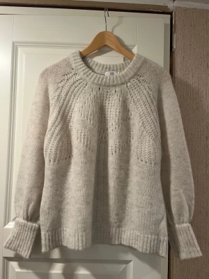 Naturvit stickad tröja från Noa Noa - Mysig mjuk naturvit stickad tröja från Noa Noa i storlek S. Tröjan har ett snyggt hålmönster upptill, breda muddar vid ärmslut och rund halsringning. Materialet är en mjuk mix av ull, alpaka och polyamid som ger en fluffig känsla. Perfekt för kyliga dagar!