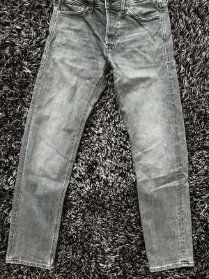 Grå jeans - Modellen heter Mike och har en Tapered fit. Det betyder att de har lite mer plats upptill men smalnar av snyggt ner mot anklarna – perfekt om man vill ha en modern look som inte känns för begränsande. Den grå färgen har en naturlig slitning som ger dem en lite lagom "insvängd" känsla direkt. • Storlek: 28/32. • Modell: Mike – Tapered fit. • Färg: Tvättad grå. • Detaljer: Klassisk femficksmodell från Jack & Jones