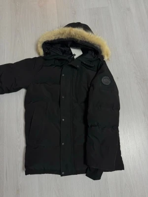 Canada Goose Carson - Säljer en svart dunjacka från Canada Goose med huva och beige päls. Jackan har lång ärm, dragkedja och knappar framtill samt ett broderat märke på ärmen. Säljs pågrund av fel storlek lapparna sitter kvar på jackan.