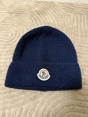 Mörkblå Moncler mössa i ull - Snygg mörkblå mössa från Moncler med klassisk ribbstickad design och uppvikt kant. Mössan har Monclers ikoniska logotyp framtill och är tillverkad i mjuk ull som håller dig varm under kyliga dagar.