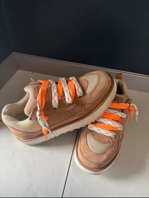 UGG Lowmel sneakers  - Säljer ett par UGG Lowmel sneakers i beigeoch nästan aldrig använda. brunt med coola orange och vita snören. Skorna har en rund tå, platt sula och är tillverkade i mocka och mesh. Perfekt för dig som gillar streetstyle och vill sticka ut med färgglada detaljer.  