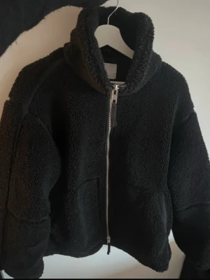 Svart fodrad teddyjacka med dragkedja - Mysig svart teddyjacka med hög krage och grov dragkedja framtill. Jackan har en loose passform och är tillverkad i fluffigt fleece-material som håller dig varm. Perfekt för kyliga dagar och har praktiska fickor på sidorna. Den är fodrad