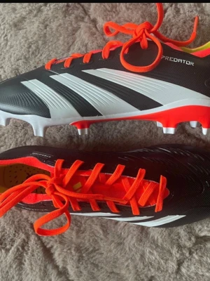 Adidas Predator fotbollsskor svart/röd - Helt nya Adidas Predator fotbollsskor med svart ovandel, vita detaljer och röda snören. Skorna har en platt sula med dobbar i vitt och rött, samt mönstrad yta för extra grepp. Insidan är gul och röd, och materialet är syntet. Står Strl 39 1/3, men skulle säga att de är lite mindre i passformen
