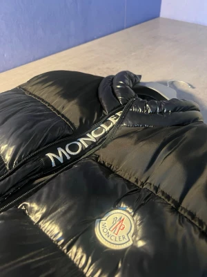 Svart dunväst från Moncler - Säljer en svart, glansig dunväst från Moncler med hög krage och tydlig logotyp på bröstet. Västen har två fickor med knappstängning och en dragkedja framtill med Moncler-text. Perfekt för att addera lite lyx till din outfit.