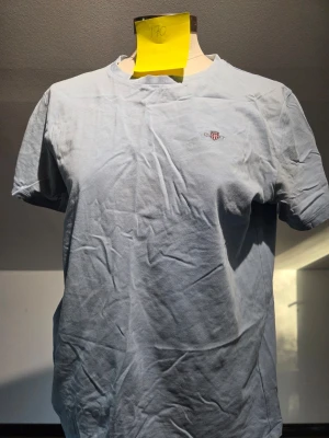Gant T-shirt - Säljer en ljusblå t-shirt från Gant med en liten broderad logga på bröstet. T-shirten har klassisk rund hals och korta ärmar, perfekt för en chill och enkel stil. Materialet är mjuk bomull som känns skönt mot huden.