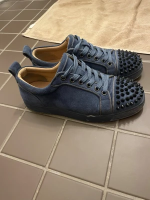 Blå Christian Louboutin sneakers med nitar - Säljer ett par blå sneakers från Christian Louboutin med coola svarta nitar på tån och klassisk röd sula. Skorna är gjorda i mocka och har snörning samt rund tå. Insidan är beige och sulan har Louboutin-loggan i rött.