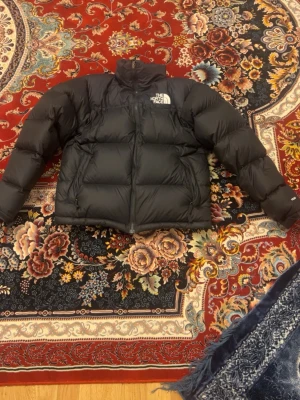  The North Face - Säljer en svart dunjacka från The North Face med klassisk puffer-design. Jackan har hög krage, dragkedja framtill och två sidofickor. Perfekt för kalla dagar och riktigt skön att bära. Loggan syns tydligt både fram och bak.