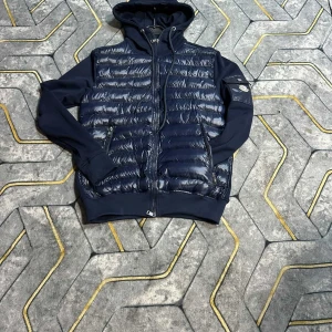 Mörkblå puffer kardigan med huva - Snygg mörkblå puffer  kardigan jacka från Moncler med mat glans quiltad framsida och mjuka ärmar. Jackan har huva med snörning, dragkedja framtill och två sidofickor med dragkedja. Perfekt för dig som vill ha en cool och sportig look.