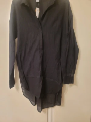 Svart randig skjorta från Lindex - Svart skjorta från Lindex i storlek S med diskreta ränder. Skjortan har lång ärm, klassisk krage och knappar framtill. Materialet är lätt och känns luftigt, perfekt för dig som gillar en chill och stilren look. Passformen är loose och skjortan är lite längre baktill.