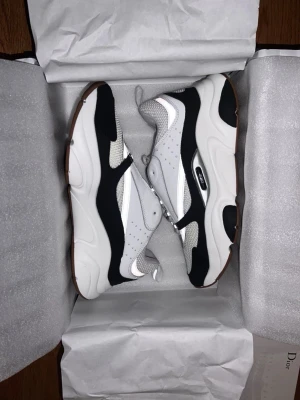 Dior B22 sneakers i vit och svart - Säljer ett par Dior B22 sneakers med chunky sula i vit, svart och grå. Skorna har meshpaneler, detaljer i skinn och syntet samt snörning. Sulan är brun undertill och har tydlig Dior-logga på sidan. Perfekt för dig som gillar exklusiva och trendiga sneakers.