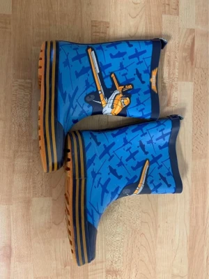 Blå stövlar med flygplansmönster - Säljer ett par coola blå stövlar med orange och mörkblå detaljer. Stövlarna har ett mönster med flygplan och en stor helikopter på sidan. Sulan är orange med ränder och grovt mönstrad för extra grepp. Perfekt för regniga dagar!