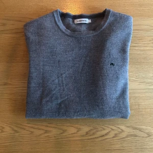 Grå merinoulltröja från J.Lindeberg - Säljer en stilren grå tröja från J.Lindeberg i 100% merinoull. Tröjan har rund halsringning, diskret logga på bröstet och ribbade muddar vid ärmslut och nederkant. Perfekt för dig som gillar klassisk och clean stil.