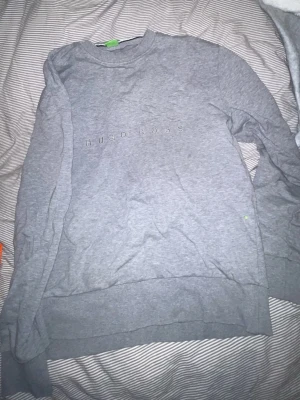 Grå sweatshirt från Hugo Boss - Säljer en stilren grå sweatshirt från Hugo Boss med broderad logga på bröstet. Tröjan har rund halsringning, ribbade muddar och är tillverkad i mjuk bomull. Perfekt för en clean och avslappnad look.