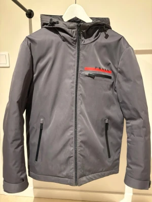 Prada vinterjacka  - Snygg grå vinterjacka från Prada med huva och svarta dragkedjor. Jackan har en röd detalj med Prada-logga på bröstet och praktiska fickor framtill. Perfekt för dig som gillar stilrena och funktionella plagg med exklusiv känsla.