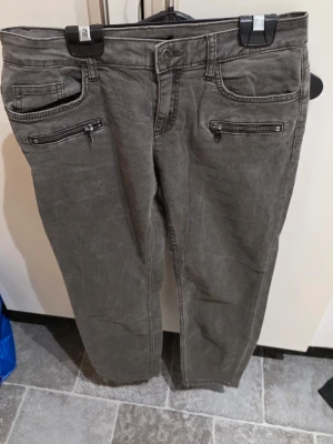 Grå jeansbyxor med dragkedjor från Lindex - Snygga grå jeansbyxor från Lindex i storlek 38. Byxorna har två framfickor med dragkedjor och klassiska bakfickor. Materialet är 96% bomull och 4% elastan vilket ger en bekväm och lätt stretchig känsla. Perfekt för dig som gillar detaljer och stilren look.