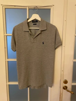 Grå pikétröja från Polo Ralph Lauren - Säljer en stilren grå pikétröja från Polo Ralph Lauren i storlek S, custom fit. Tröjan har klassisk krage, korta ärmar och två knappar framtill. Materialet är mjukt och känns som bomull. Diskret logga broderad på bröstet i mörkblått.