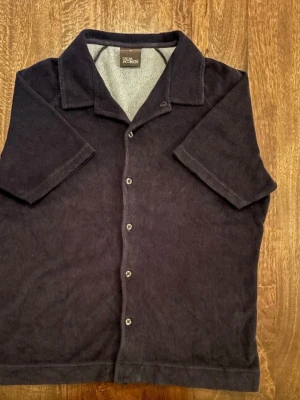 Mörk blå poloshirt/frotte från Oscar Jacobson - Mörk blå poloshirt/frotte från Oscar Jacobson i mjuk frotté med klassisk krage och korta ärmar. Skjortan har knappar framtill och en loose passform som ger en chill vibe. Perfekt för dig som gillar stilren design och bekvämt material.