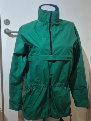 Grön vindjacka  - Grön vindjacka med hög krage och half zip framtill. Jackan har en stor ficka med dragkedja på magen och snörning i midjan för justerbar passform. Materialet är lätt och syntetiskt, perfekt för utomhusaktiviteter. Insidan har en blå detalj vid kragen.💚💙🛍 storlek M