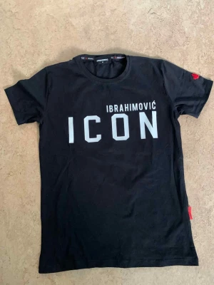 Svart ICON t-shirt från Dsquared2 - Svart t-shirt från Dsquared2 med trycket 'IBRAHIMOVIĆ ICON' på framsidan och en stor svartvit bild med DSQUARED2-logga på ryggen. Rund hals och korta ärmar, tillverkad i mjuk bomull. Röd detalj på ärmen och etikett i sidan ger extra edge.