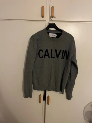 Grå sweatshirt från Calvin Klein Jeans - Snygg grå sweatshirt från Calvin Klein Jeans med stor svart CALVIN-text på bröstet. Tröjan har rund hals, ribbade muddar vid ärmslut och nederkant samt långärmad passform. Perfekt för en chill och stilren look.