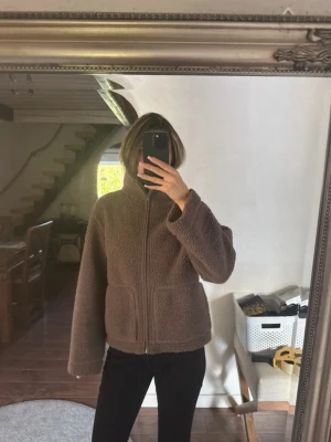 Brun teddyjacka med dragkedja - Mysig brun teddyjacka med hög krage och två stora fickor framtill. Jackan har en rak och loose passform med långärmade ärmar och stängs med dragkedja. Perfekt för kyliga dagar när du vill vara både bekväm och snygg.