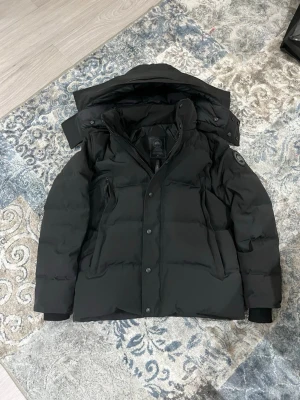 Canada goose | Wyndham edition - Svart dunjacka från Canada Goose med avtagbar huva och klassisk logotyp på ärmen. Jackan har dragkedja och knappar framtill, samt flera fickor. Medföljer en pälsdetalj till huvan. Perfekt för kalla vinterdagar och har en clean, stilren look.