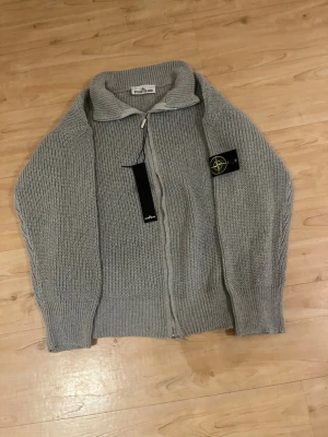 Grå stickad kofta från Stone Island - Säljer en grå stickad kofta från Stone Island med dragkedja framtill och ribbade muddar. Koftan har lång ärm, klassisk krage och den ikoniska Stone Island-patchen på vänster ärm. Perfekt för dig som gillar stilrena och bekväma plagg.