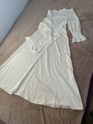 Vit långklänning med volangärmar - Elegant vit långklänning i satinliknande material med markerad midja och knytband. Klänningen har långa ärmar med volangdetaljer och silvriga knappar vid ärmsluten. Rundad halsringning och diskret dragkedja bak för enkel påklädning.