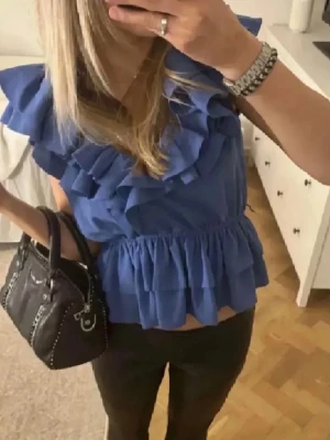 Blå volangblus med ärmlös design - Säljer en blå blus med flera volangdetaljer både vid axlarna och längs nederkanten. Toppen är ärmlös och har en elastisk midja som ger en snygg passform. Perfekt för dig som gillar feminina detaljer och en luftig känsla. Från hm 