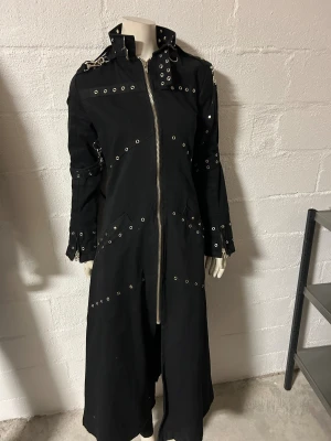 Svart lång rock med nitar och dragkedjor stl S. - Säljer en svart lång rock med coola metalldetaljer, nitar och öljettband över hela jackan. Jackan har hög krage med spänne, långa ärmar med dragkedjor och flera ringar och kedjor för en edgy look. Materialet är kraftig bomull och passformen är normal. Storlek S. Helt ny. Märke Lucifer.