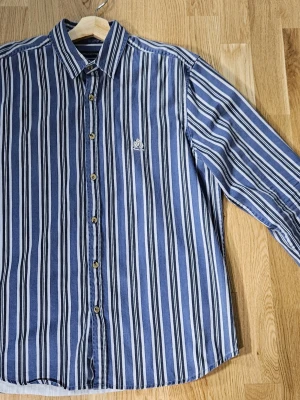 Blåvit randig skjorta Urban Outfitters - Snygg blå och vit randig skjorta från Urban Outfitters med broderad logga på bröstet. Skjortan har klassisk krage, långa ärmar och knappar framtill. Tillverkad i mjuk bomull som känns skön mot huden. Perfekt för dig som gillar stilrena och fräscha plagg.