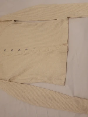 Beige ribbad kofta från Lindex XS - Säljer en beige ribbad kofta från Lindex i storlek XS. Koftan har långa ärmar och är tillverkad i mjuk bomull. Den har snygga tryckknappar i silver längs framsidan och ett stilrent, enkelt uttryck. Perfekt för lager-på-lager under höst och vår.