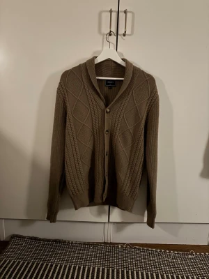 Beige stickad kofta - Snygg beige stickad kofta från Baker's med grovt kabelmönster och rutstruktur på framsidan. Koftan har ribbade muddar, v-ringad krage och knappar i guldton. Perfekt att slänga på sig när det är lite kyligt ute. Storlek L men sitter som storlek M. Modellen i bilden är 180 och väger 72 kg.