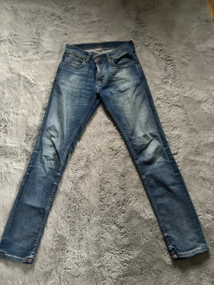 Grim Tim ”worn in broken” från Nudie - Säljer ett par Grim Tim  från Nudie  med klassisk femficksdesign och snygga slitningar. Jeansen har orangea sömmar och Nudie Jeans typiska broderi på bakfickan. Perfekt passform och cool tvätt för dig som gillar streetstil. Storlek 30x32